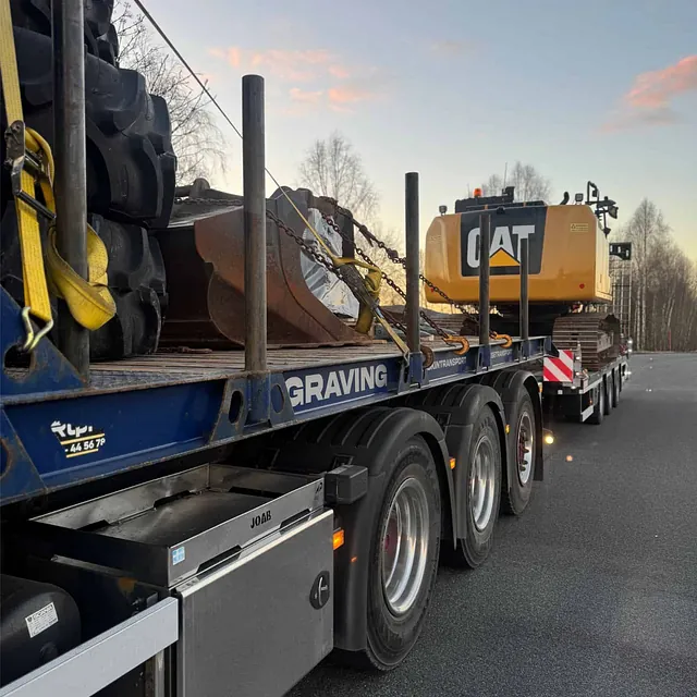 Lastebil med gravemaskin og utstyr på tilhenger, parkert ved veikant i solnedgang.