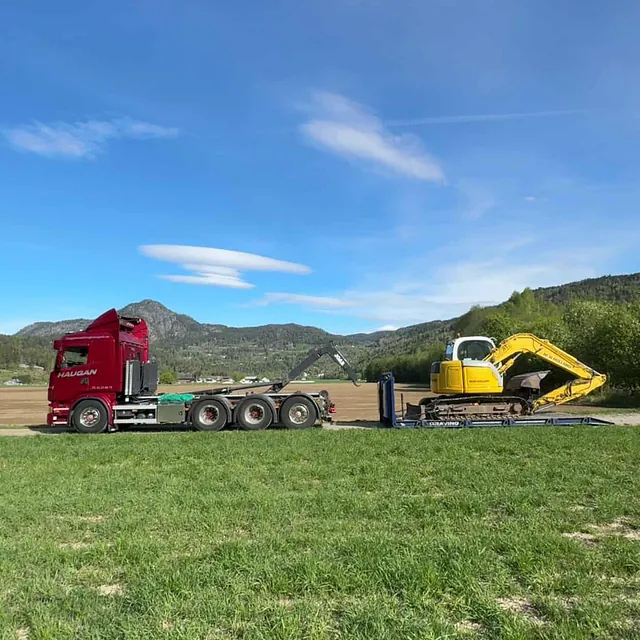 Rød Haugan-lastebil med krokhenger og gravemaskin i ferd med opplasting på jorde omgitt av fjell.