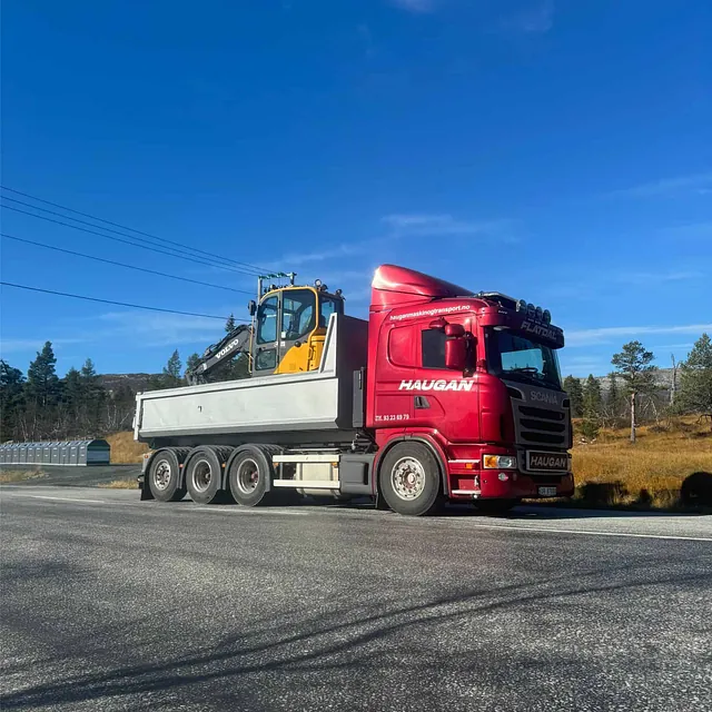 Rød Haugan-lastebil transporterer liten gul gravemaskin i tippkasse, parkert langs vei.