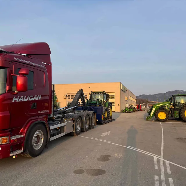 Rød Haugan-lastebil og flere traktorer parkert ved industribygg med fjell i bakgrunnen.