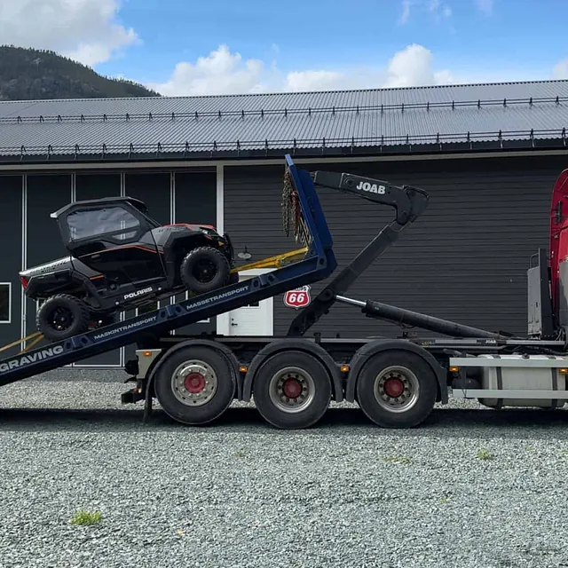 Sort Polaris ATV lastet på krokhenger med JOAB-arm, parkert på grusplass foran mørkt bygg.