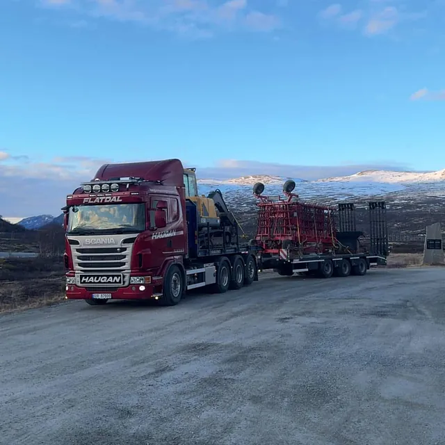 Rød Haugan-lastebil med anleggsmaskin og landbruksredskap på spesialhenger, parkert ved fjellområde.
