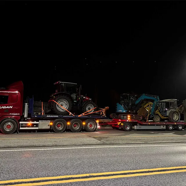 Rød Haugan-lastebil frakter tre maskiner: traktor og to gravemaskiner, under gatebelysning om natta.