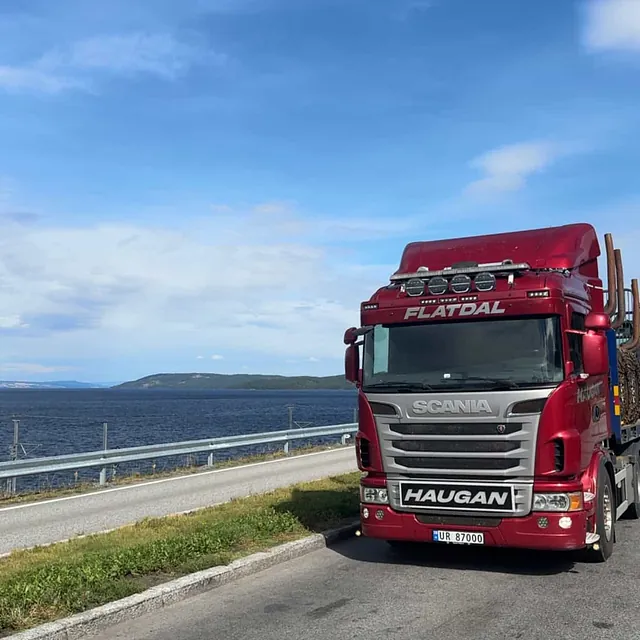 Rød Haugan-lastebil parkert ved utsiktsplass med hav og fjell i bakgrunnen, blå himmel.
