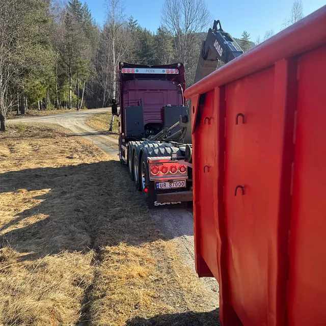 Rød Haugan-lastebil sett bakfra, klar til å hente rød container med JOAB-løftearm, omgitt av skog.