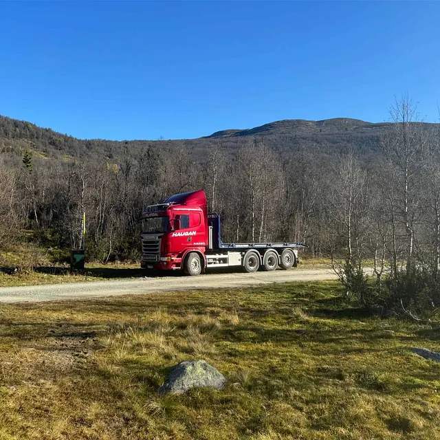 Rød Haugan-lastebil uten last parkert på grusvei i fjellområde med blå himmel.