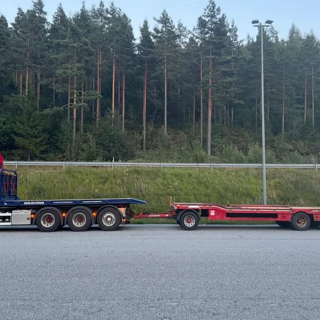 Tom rød Haugan-lastebil med lavbygget maskinhenger parkert ved veiskulder foran grønn skog.